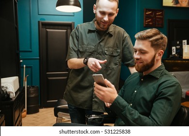 Cheerful Young Man Getting Haircut By Hairdresser While Sitting In Chair. Holding Phone.