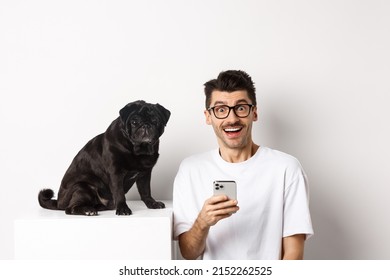 Cheerful Young Man Hipster Staring At Camera, Sitting With Cute Black Pug Dog And Using Mobile Phone, Standing Over White Background