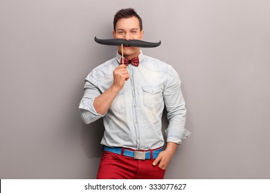 Cheerful young man holding a huge fake moustache below his nose and looking at the camera