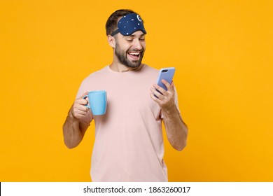 Cheerful young man in pajamas home wear sleep mask hold cup coffee tea drinking using mobile cell phone rest at home isolated on yellow background studio portrait. Relax good mood lifestyle concept - Powered by Shutterstock