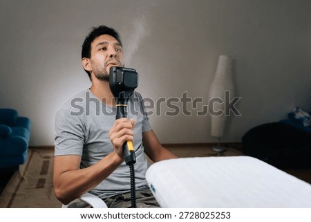 Cheerful young man playfully using steam cleaner as microphone, blowing visible steam plumes enjoying break from doing household work, making mundane tasks entertaining.