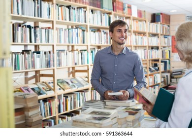 Cheerful Young Man Seller Helping Customer In Book Store