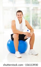 Cheerful Young Man Sitting On Gym Ball, Holding Water Bottle