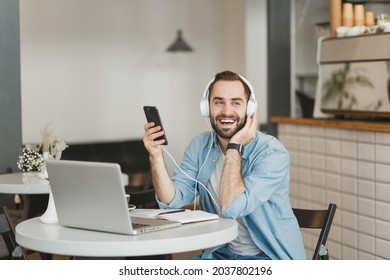 Cheerful young man sitting at table in coffee shop cafe restaurant indoors working studying on laptop pc computer listening music with headphones cell phone. Freelance mobile office business concept