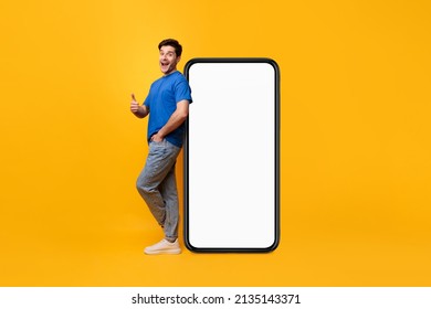 Cheerful Young Man Standing Leaning On Big Huge New Cell Phone With White Empty Screen And Showing Thumb Up Gesture, Mockup, Casual Guy Recommend Cool Mobile App, Bright Yellow Background, Full Length