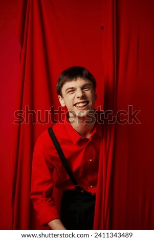 cheerful young man in suspenders smiling while hiding behind red vibrant curtain, merriment
