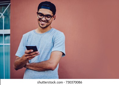 Cheerful young man in trendy baseball cap watching funny video on mobile phone using 4G connection, smiling hipster guy reading good news from networks standing on wall promotional background