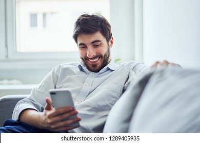 Cheerful young man using phone while relaxing on sofa after work