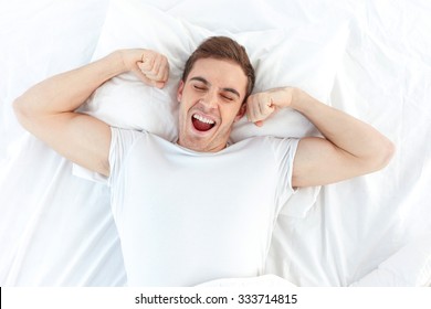 Cheerful young man is waking up after sleeping in the morning. He is yawing and stretching his arms up. His eyes are closed with relaxation. He is lying in the bed