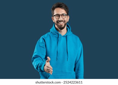 A cheerful young man wearing a blue hoodie offers a handshake with a warm smile, creating an inviting and friendly atmosphere suitable for personal or professional interactions. - Powered by Shutterstock