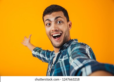 Cheerful young man wearing plaid shirt standing isolated over orange background, taking a selfie