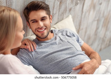 Cheerful Young Man And Woman Are Lying And Talking In Bed. They Are Hugging With Love And Smiling