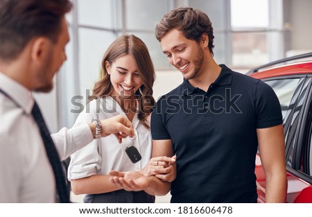 Cheerful young man and woman receiving keys of new automobile from professional dealer in car salon