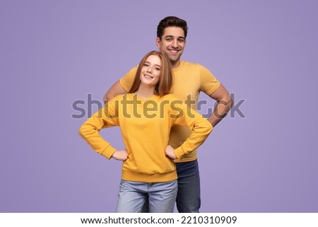Cheerful young man and woman in similar clothes holding hands on waist and looking at camera with smile, while being part of team together against violet background