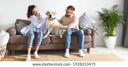 Cheerful young man and woman sitting on sofa and hugging adorable Labrador retriever dog while enjoying free time together at home