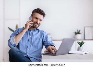 Cheerful Young Man Working Remotely From Home Office With Cellphone And Laptop, Sitting At Desk In Room, Copy Space - Shutterstock ID 1805975317