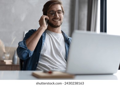 Cheerful Young Man Working Remotely From Home Office With Cellphone And Laptop, Sitting At Desk In Room, Copy Space - Powered by Shutterstock