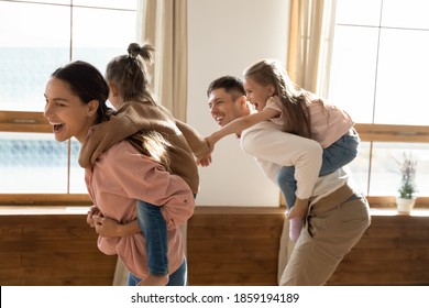 Cheerful Young Married Couple Piggyback Riding Small Daughters Play Together Active Games Running In Living Room Catching Each Other Having Fun In Modern Living Room. Family Playtime With Kids Concept