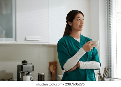 Cheerful young medical nurse in uniform drinking cup of morning coffee in kitchen - Powered by Shutterstock