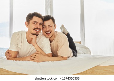 Cheerful young men smiling for camera and hugging while relaxing on comfortable bed against window in morning at home
