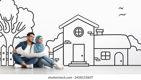 Cheerful young middle eastern couple sitting by white wall with a house outline, dreaming of a home. Real estate, mortgage concept - Powered by Shutterstock