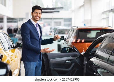 Cheerful Young Middle Eastern Man In Formal Outwear Sales Manager Showing Nice Sports Car, Open Black Auto Door And Smiling, Luxury Automobile Dealership Salon Interior, Copy Space