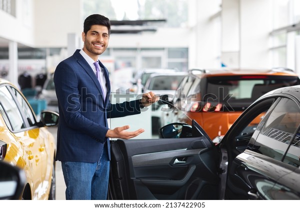Cheerful\
young middle eastern man in formal outwear sales manager showing\
nice sports car, open black auto door and smiling, luxury\
automobile dealership salon interior, copy\
space