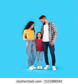 Cheerful Young Middle Eastern Parents Hugging Their Lovely Daughter Posing On Blue Background In Studio. Arabic Family Embracing Together. Square Shot, Full Length