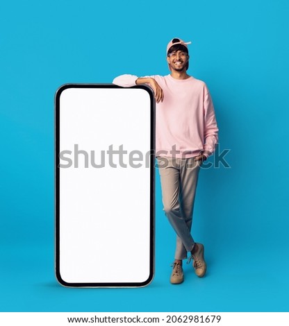 Cheerful young middle-eastern man standing by huge vertical brand new cellphone with empty screen and smiling at camera, mockup, handsome guy enjoying newest entertaining mobile app