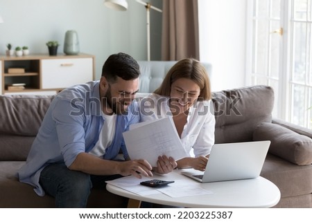 Cheerful young millennial husband and wife doing domestic paperwork, accounting job, reviewing paper bills, receipts at laptop computer, using calculator, paying mortgage, rent fees on internet