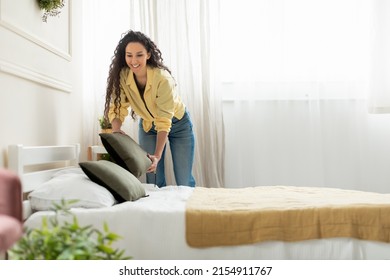 Cheerful young millennial lady making bed at home, holding pillow, doing housework in the morning. Positive stylish woman cleaning up her bedroom, everyday routine concept, free copy space - Shutterstock ID 2154911767