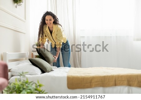 Cheerful young millennial lady making bed at home, holding pillow, doing housework in the morning. Positive stylish woman cleaning up her bedroom, everyday routine concept, free copy space