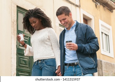 Cheerful young mix raced couple walking outdoors and minding their steps. Happy Caucasian man and black woman with takeaway coffee holding hands and looking down. Watching steps concept - Powered by Shutterstock