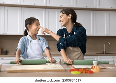 Cheerful young mom and cute helper daughter child cooking and playing. Happy mum touching face of kid with finger, leaving floury spot on nose, laughing, teaching girl to bake - Powered by Shutterstock