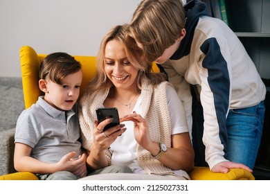 Cheerful Young Mother With Childs Having Fun Using Smartphone At Home Sitting Hugs In Cozy Yellow Armchair. Pretty Mom With Little Sons Kid In Interior Of Cozy Living Room Looking At Screen Of Phone.