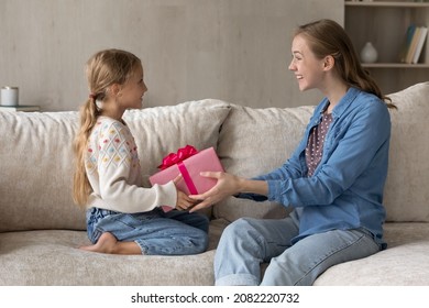 Cheerful young mother giving wrapped gift box to excited small kid daughter, congratulating with happy birthday or making surprise present, sitting together on comfortable sofa in living room.