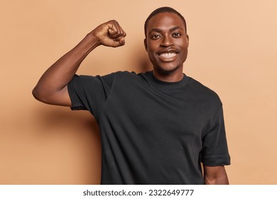 Cheerful young muscular athletic man dressed in casual tshirt proudly flexes biceps after intense training session at gym demonstrates results of his hard work and dedication isolated on brown wall - Powered by Shutterstock