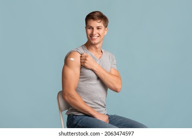 Cheerful young muscular attractive european guy showing hand with band aid after injection, isolated on blue background. New normal, successful covid-19 vaccination, health care and immunization