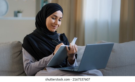 Cheerful young muslim arabian lady young woman sit on cozy sofa female freelancer writer take notes in diary noting information write book daily tasks secrets ideas in personal organizer using laptop