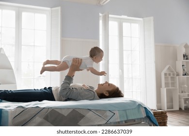 Cheerful young new mom holding kid, lifting sweet positive baby up in air, lying on bed, smiling, laughing, playing airplane with infant child, enjoying leisure time, motherhood