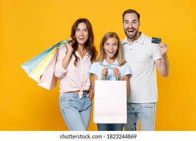 Cheerful Young Parents Mom Dad With Child Kid Daughter Teen Girl In Basic T-shirts Hold Package Bags With Purchases Credit Bank Card Isolated On Yellow Background Studio Portrait. Family Day Concept