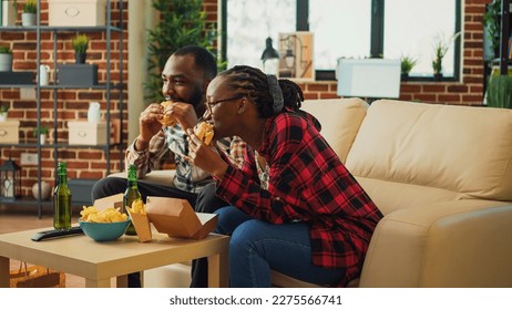 Cheerful young people eating hamburgers and fries in living room, watching favorite film on tv and drinking alcohol. Boyfriend and girlfriend serving fast food order from takeout place. - Powered by Shutterstock