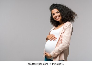 cheerful young pregnant african american woman touching belly on grey 