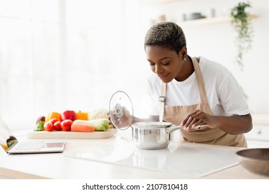 Cheerful Young Pretty Black Female In Apron Prepare Lunch And Smell Dish In Minimalist Kitchen Interior. Cooking Dinner For Family At New Recipe At Home. Food Blog During Covid-19 Pandemic, Free Space