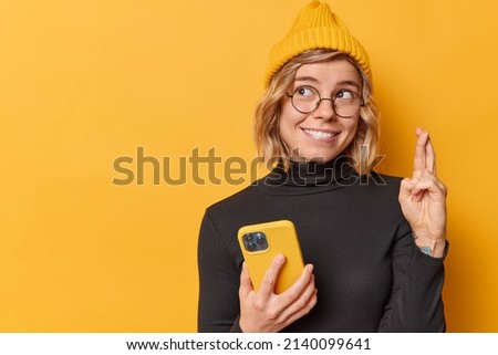 Cheerful young pretty woman keeps fingers crossed makes wish holds smartphone waits for dreams come true wears hat black turtleneck round spectacles isolated over yellow background copy space