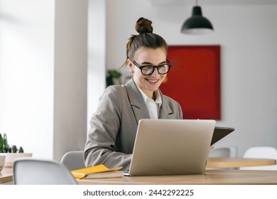 A cheerful young professional immersed in her laptop, sporting trendy glasses and a smart houndstooth blazer, in an office with a touch of art.

 - Powered by Shutterstock