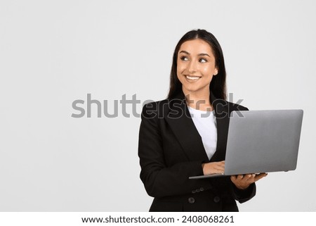 Cheerful young professional woman in business attire holding an open laptop, looking to the side at copy space with smile, on simple grey backdrop