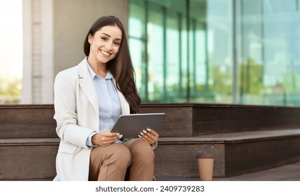 Cheerful young professional woman sitting on bench with digital tablet, smartwatch on wrist, next to coffee cup, smiling and looking at camera with modern glass building in the background - Powered by Shutterstock