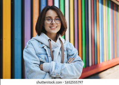 Cheerful young queer girl in glasses, denim jacket, standing near rainbow wall, cross hands over chest and smiling camera happy, lifestyle, urban life and generation concept