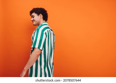 Cheerful Young Queer Man Standing Against An Orange Background. Side View Of A Happy Young Generation Z Hipster Looking Confident While Standing Alone In A Studio.